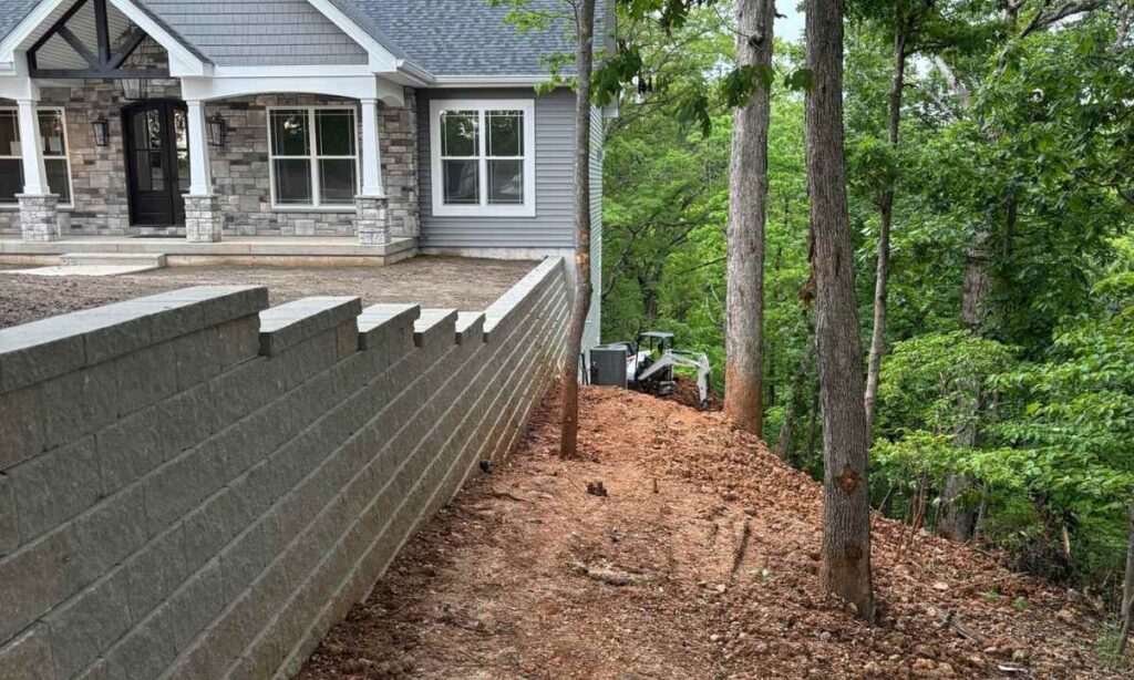 Sloped backyard excavation with retaining wall installation in progress near trees, showcasing proper seasonal timing for wall builds in St. Louis