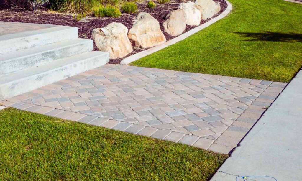 Brick paver pathway in a landscaped St. Louis front yard connecting concrete steps to a sidewalk, bordered by grass and large decorative stones.