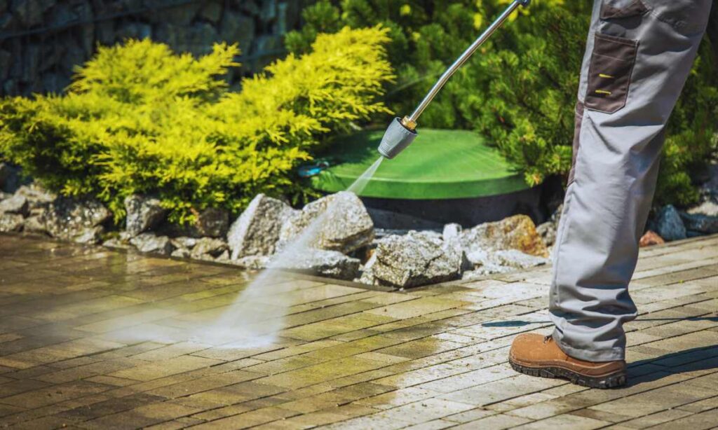 Worker in boots pressure washing a stone paver patio near landscaping rocks and shrubs in St. Louis
