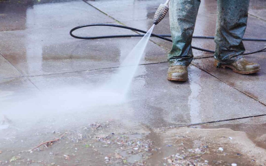 worker pressure washing a concrete paver surface in St. Louis, showing water spray and protective boots for safe outdoor cleaning.