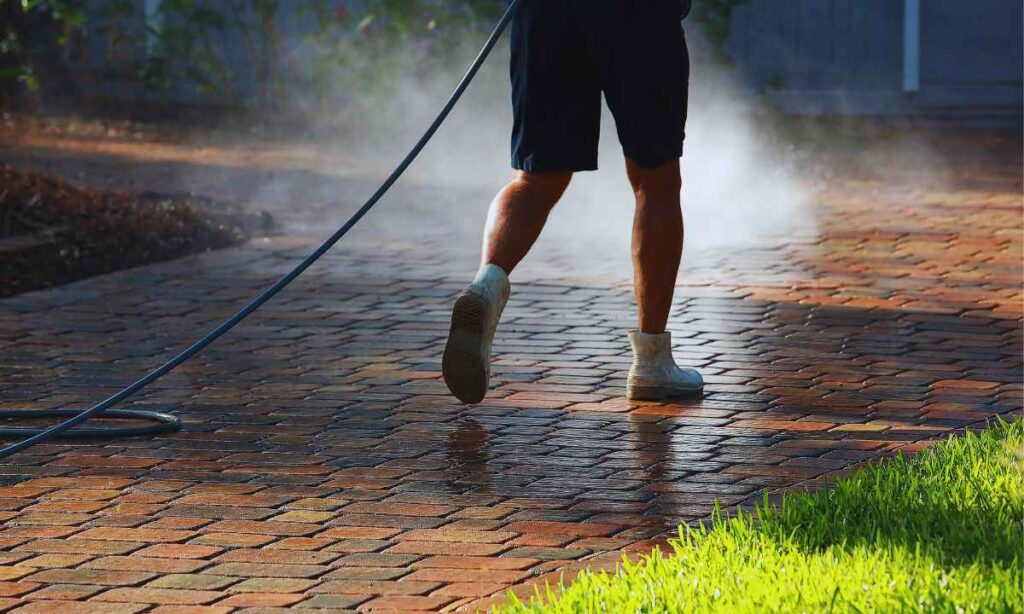 A professional worker pressure-washes a brick paver driveway in St. Louis, removing dirt and buildup to prepare the surface for sealing. Sunlight highlights clean, wet pavers and nearby green grass, showing the results of proper hardscape maintenance.