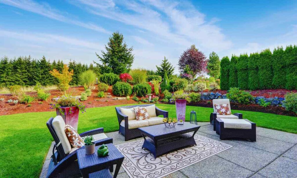 Modern St. Louis backyard patio featuring large square concrete pavers, woven outdoor lounge furniture, a patterned rug, and vibrant garden landscaping with evergreens and flowering shrubs—ideal for creating a relaxing and stylish outdoor living area.