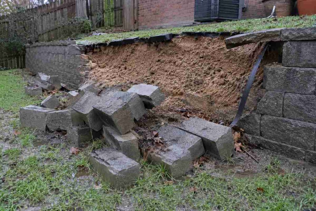 Partially collapsed residential retaining wall with displaced blocks and exposed soil, showing conditions to document before repair