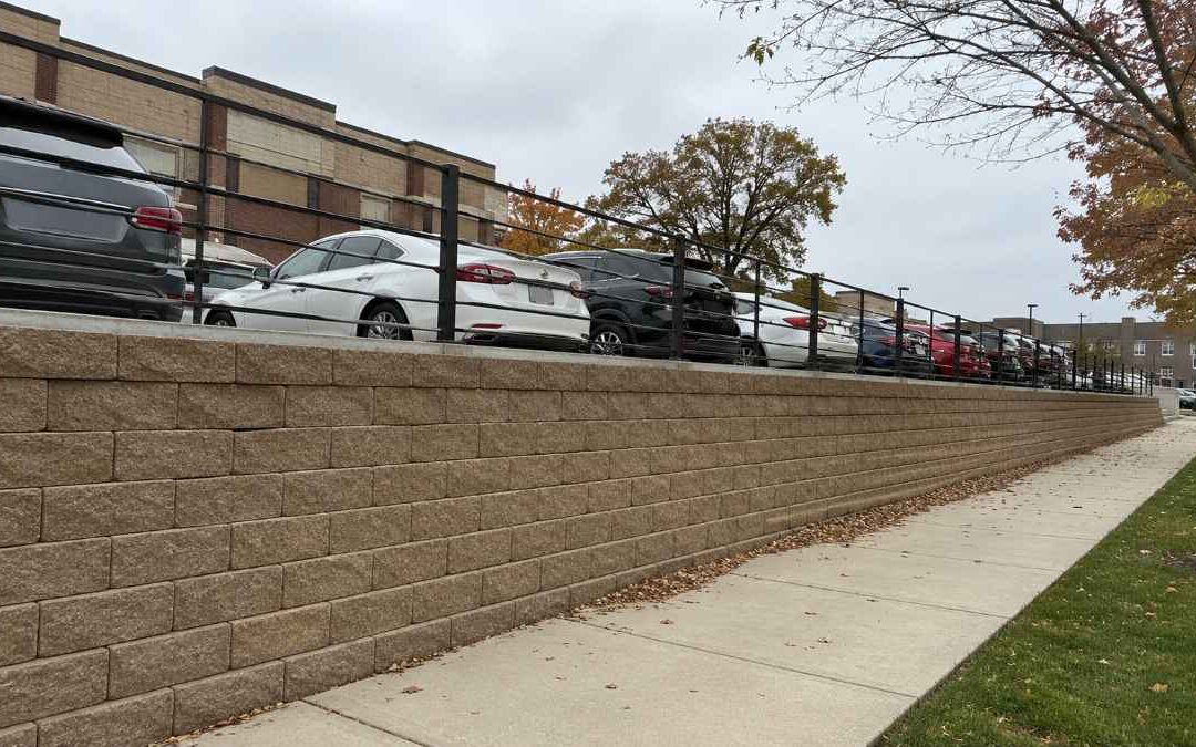 Engineered segmental retaining wall holding back a raised commercial parking lot with cars above and a guard rail along the top edge.